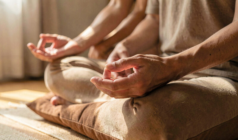 Close view of hands resting in meditation posture on folded legs, warm indoor light, simple cushion.
