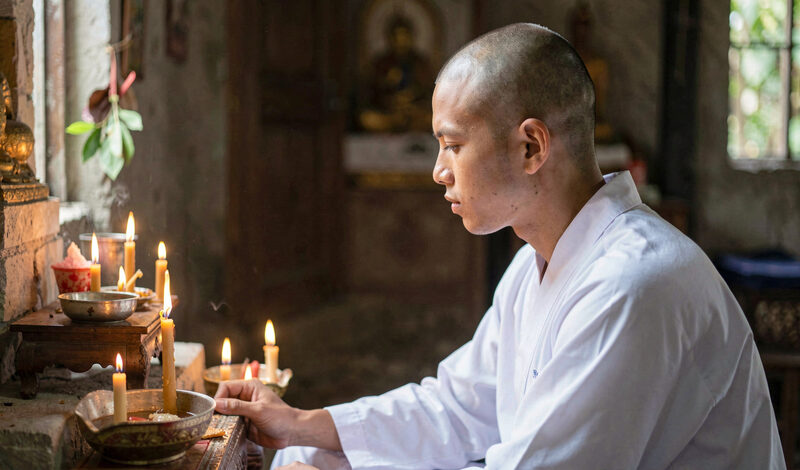 A man with freshly shaved head wearing white ordination robes sitting before a small shrine.